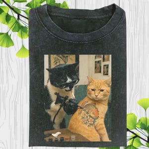 May include: Dark gray t-shirt featuring a humorous illustration of two cats. One cat, wearing glasses, is tattooing a koi fish design on the other cat's shoulder. The tattoo includes flowers and a koi fish. The image is set against a neutral background.