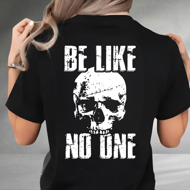 May include: Black t-shirt with a white skull graphic and the text "BE LIKE NO ONE". The skull design is detailed with shading and texture. The text is in a bold, blocky font, creating a striking contrast against the black fabric.