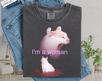 May include: Dark gray t-shirt with a graphic of a white cat with the text "I'm a woman" in pink. The shirt is folded on top of blue denim jeans.