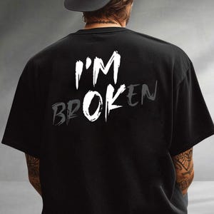 May include: Black t-shirt with the words "I'M BROKEN" printed on the back in white and gray. The text is stylized with a distressed font, creating a bold statement. The shirt is a casual wear item.