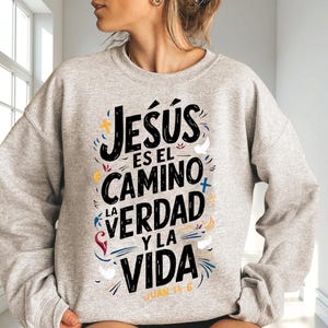 May include: A light gray sweatshirt with the text "Jesús es el Camino, la Verdad y la Vida" in black, with colorful accents. The text is in a stylized font, with the Bible verse "Juan 14:6" at the bottom. The sweatshirt is a casual style.