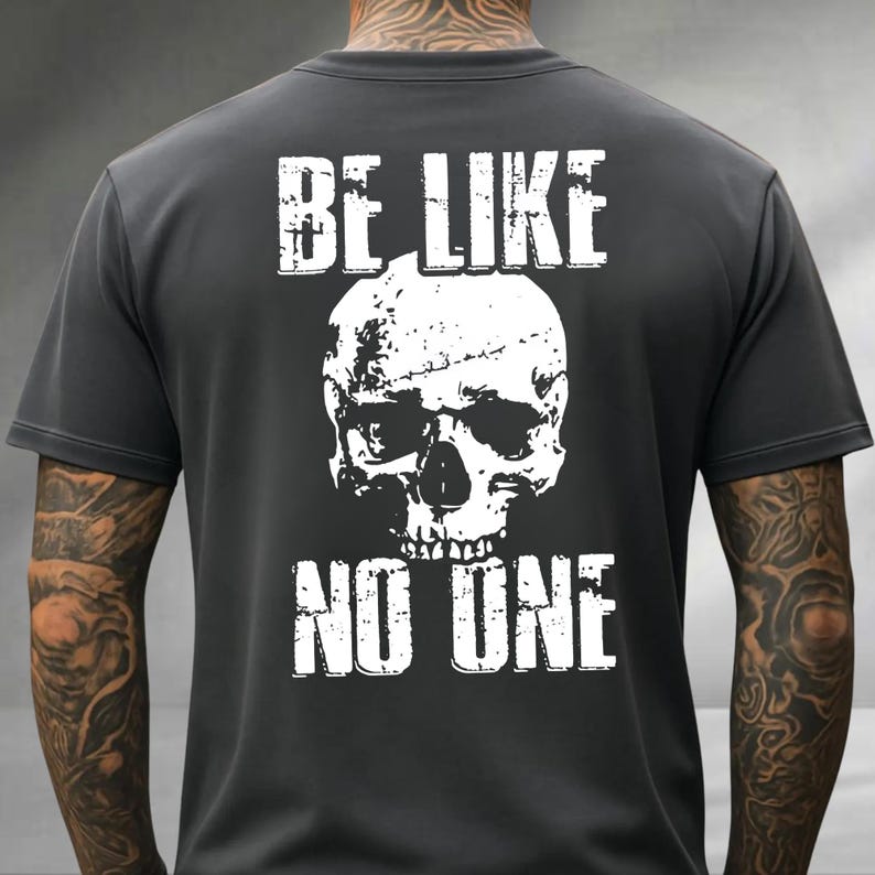 May include: Dark gray t-shirt with a white skull graphic and the text "BE LIKE NO ONE". The design is distressed, giving it a vintage look. The shirt is made of a soft material.