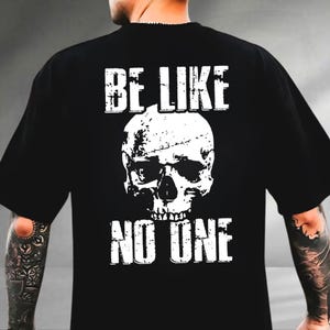 May include: Black t-shirt featuring a distressed white skull graphic. The text "BE LIKE" is above the skull, and "NO ONE" is below it. The shirt has a classic crew neck and short sleeves.