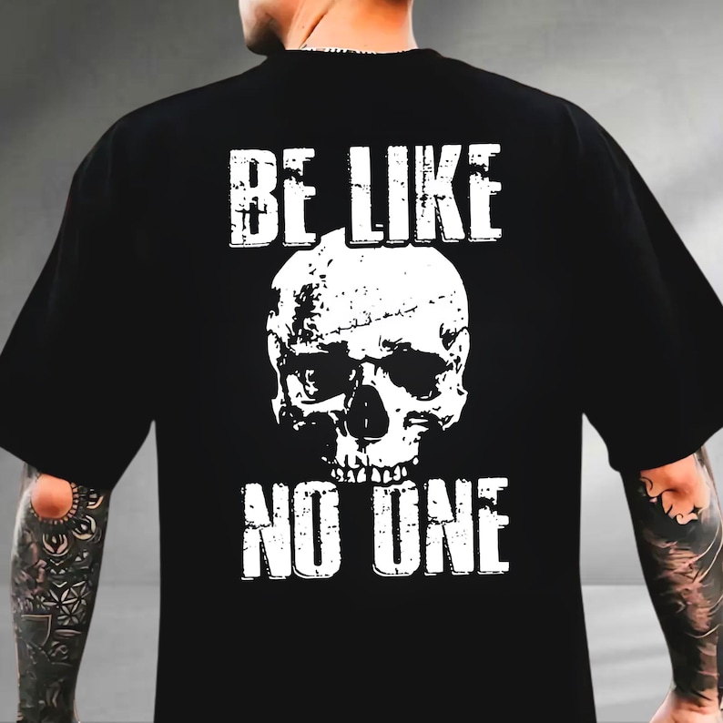 May include: Black t-shirt featuring a distressed white skull graphic. The text "BE LIKE" is above the skull, and "NO ONE" is below it. The shirt has a classic crew neck and short sleeves.