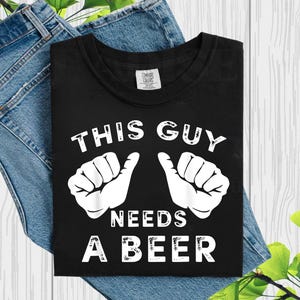 May include: Black t-shirt with the text "THIS GUY NEEDS A BEER" in white, along with two thumbs-up hand illustrations. The shirt is displayed on a light wood surface with denim jeans.