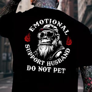 May include: Black t-shirt featuring a gorilla wearing sunglasses and a cap, encircled by the words "EMOTIONAL SUPPORT HUSBAND DO NOT PET" in white. Red flame graphics flank the gorilla image. The design is centered on the back of the shirt.