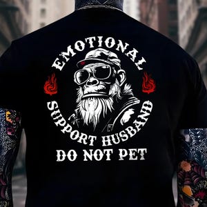 May include: Black t-shirt featuring a gorilla wearing sunglasses and a cap, with the text "EMOTIONAL SUPPORT HUSBAND DO NOT PET" in a circular design. The design includes flames and is printed in white.