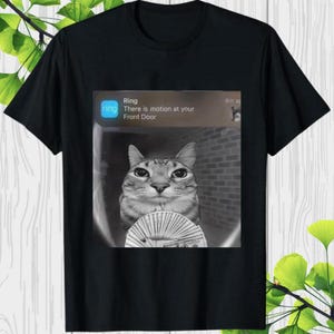 May include: Black t-shirt featuring a grayscale image of a cat holding money, with a Ring notification that reads "There is motion at your Front Door." The cat is the central focus, with a brick wall background.