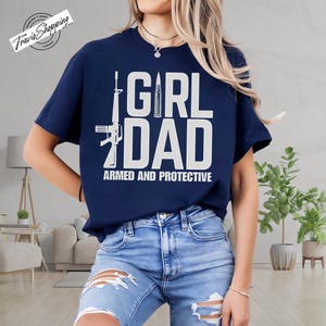 May include: Navy blue t-shirt with the text "GIRL DAD ARMED AND PROTECTIVE" in white. The design includes illustrations of a rifle and a bullet. The shirt is a short-sleeved crew neck.