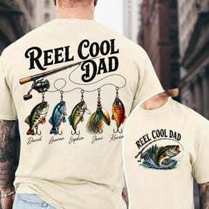 A person wearing a white t-shirt with a design featuring a fishing rod and various fish illustrations, along with the text "Reel Cool Dad" and names of the people involved in the fishing activity.