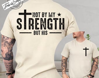 May include: A beige t-shirt with the text "NOT BY MY STRENGTH BUT HIS" in black, along with a cross and star design. A smaller image shows the same shirt with a cross on the chest.