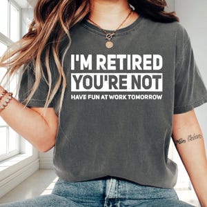 May include: Dark gray t-shirt with the text "I'M RETIRED YOU'RE NOT HAVE FUN AT WORK TOMORROW" in white block letters. The shirt is a crew neck and has short sleeves. The shirt is worn by a person.