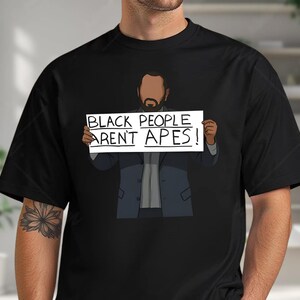 May include: Black t-shirt featuring an illustration of a person holding a sign that reads "BLACK PEOPLE AREN'T APES!". The person is wearing a suit jacket. The shirt is black.