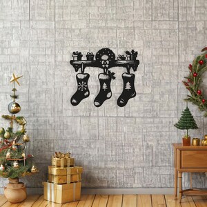 Puede incluir: Porta calcetines de Navidad de metal negro con tres calcetines colgando. Los calcetines están decorados con copos de nieve y un árbol de Navidad. El soporte está montado en una pared de ladrillo blanco.