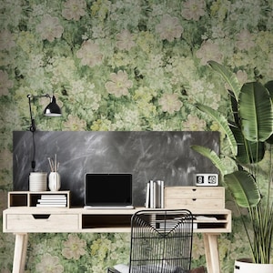 May include: A light wood desk with a black metal chair and a laptop. The desk is in front of a floral wallpaper with green and pink flowers. A large potted plant is to the right of the desk. A black lamp is mounted on the wall.