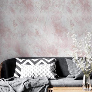 May include: A contemporary living room setting with a grey sofa, patterned cushions, and a grey throw. The backdrop is a textured wall with pink and white paint strokes. A vase of white flowers is on a wooden table.