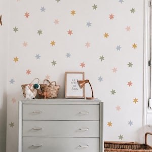 Puede incluir: Una cómoda gris claro de cuatro cajones con tiradores plateados, situada contra una pared decorada con pegatinas de estrellas en tonos apagados de rosa, azul, amarillo y verde. Un cartel enmarcado dice LET THEM BE little junto a una lámpara.