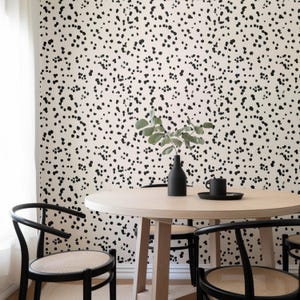 May include: A dining room scene featuring a round wooden table and black chairs with woven seats. A black vase with greenery and a black mug on a small tray sit on the table. The wall is covered in a cream-colored wallpaper with a black dot pattern.