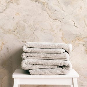 May include: A stack of four folded, light gray bath towels rests on a small, white, square stool. The background features a marble-like wall with beige and brown veining, creating a neutral and spa-like aesthetic. The towels appear soft and plush.