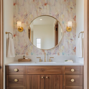 May include: A contemporary bathroom vanity featuring a circular mirror and gold fittings. The vanity unit is crafted from dark wood with gold handles. The wall behind the mirror is adorned with colourful abstract wallpaper. Two sconces are positioned either side of the mirror, with towels on gold towel rails.