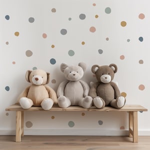 May include: Three plush teddy bears, one brown, one beige, and one gray, are sitting on a wooden bench in front of a white wall with colorful polka dot stickers.