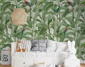 Puede incluir: Un dormitorio infantil con una cama blanca, juguetes y papel pintado de hojas tropicales verdes con pájaros de colores. Una lámpara colgante tejida cuelga del techo. Una almohada gris dice "LOVE YOU TO THE MOON AND BACK".