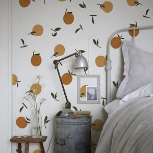 Könnte beinhalten: Schlafzimmer-Interieur mit einer weißen Wand, die mit Orangen- und grünen Blattaufklebern verziert ist. Es gibt ein Metallbettgestell, eine Vintage-Lampe und einen kleinen Holzschemel mit einer Blumenvase. Die Einrichtung wirkt rustikal und gemütlich.