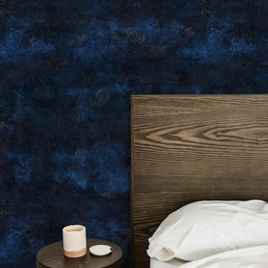 Puede incluir: Papel pintado abstracto azul oscuro y dorado con una cabecera de madera y una pequeña mesa auxiliar redonda de color marrón oscuro. Una taza blanca con base rosa y una pequeña tapa blanca sobre la mesa. Ropa de cama blanca visible.