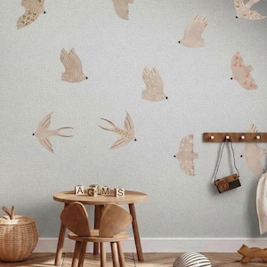 Könnte beinhalten: Eine hellblaue Wand mit mehreren beigefarbenen Vogelaufklebern. Vor der Wand befindet sich ein kleiner Tisch und Stuhl aus Holz. Ein geflochtener Korb und Holzblöcke mit der Aufschrift "AGLMS" stehen auf dem Tisch. Eine Holzgarderobe mit Kamera und Tasche hängt an der Wand.