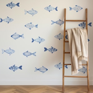 Pode incluir: Uma parede branca com um papel de parede de padrão de peixe azul e branco. Uma escada de madeira com um cobertor bege drapeado sobre ela está encostada na parede.
