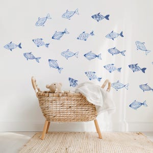 May include: A white wall with blue watercolor fish decals. A wicker bassinet with a white blanket sits in front of the wall.
