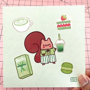May include: A square illustration featuring a cartoon squirrel surrounded by food and drink items. The squirrel is wearing a green shirt and striped pants. Items include matcha, boba tea, cake, macarons, and matcha biscuit sticks.