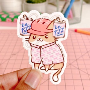 May include: A sticker featuring a cartoon cat wearing a pink hat and a pink patterned shirt. The cat is holding two cans of "Le Fizz" soda. The sticker has a white border and is held by a hand. The background is a pink cutting mat.