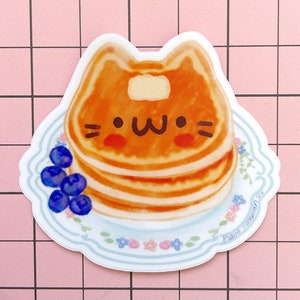 Puede incluir: Una pegatina de una pila de panqueques con forma de gato con una cara linda. Los panqueques est&aacute;n en un plato blanco con un patr&oacute;n floral azul y blanco. Hay ar&aacute;ndanos en el plato. La pegatina tiene el texto "brave fashion" en la esquina inferior derecha.