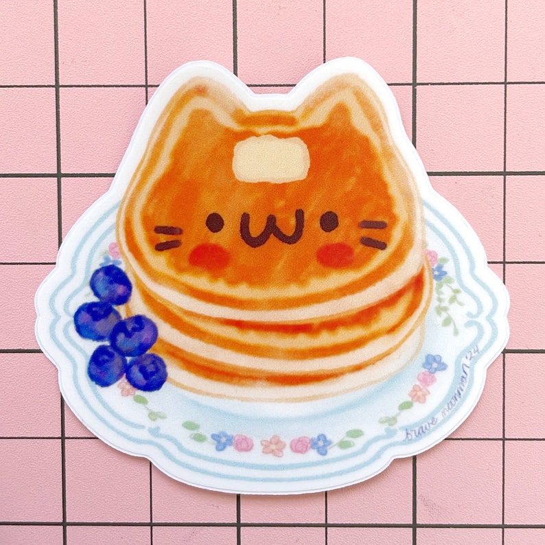 Puede incluir: Una pegatina de una pila de panqueques con forma de gato con una cara linda. Los panqueques est&aacute;n en un plato blanco con un patr&oacute;n floral azul y blanco. Hay ar&aacute;ndanos en el plato. La pegatina tiene el texto "brave fashion" en la esquina inferior derecha.
