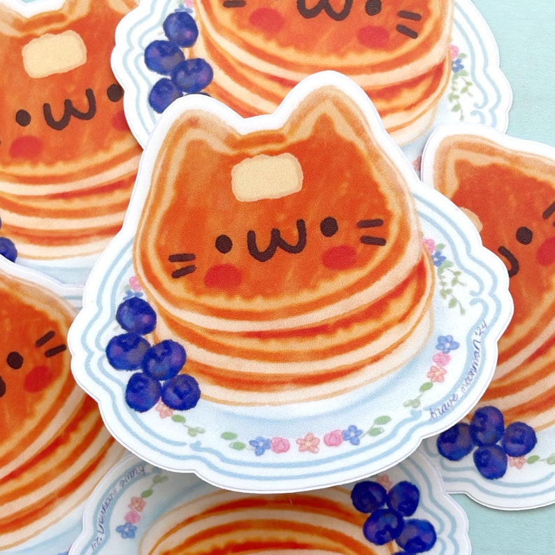 Puede incluir: Una pila de panqueques con forma de gato lindo con una cara sonriente, cubierta con mantequilla y rodeada de ar&aacute;ndanos en un plato azul y blanco con un borde floral. La pegatina dice "brave cinnamon"
