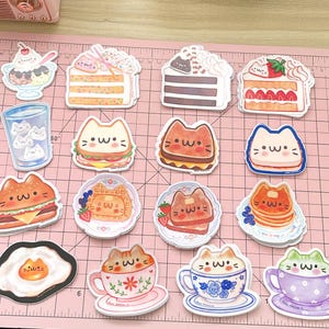 May include: A set of 14 colorful stickers featuring cute cartoon cats enjoying various foods and drinks. The stickers include a cat eating a hamburger, a cat drinking coffee, a cat eating waffles, and a cat eating a slice of cake.