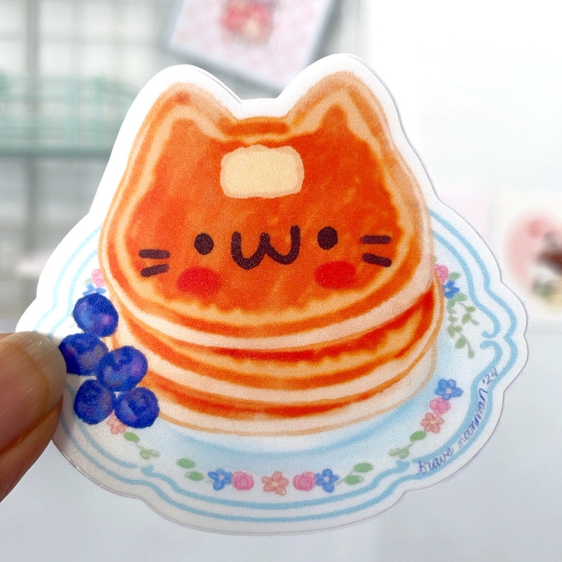 Puede incluir: Una pegatina con un panqueque con forma de gato de dibujos animados con una cara feliz, sentado en un plato azul y blanco con ar&aacute;ndanos y flores. El texto "brave mammal" est&aacute; impreso en el plato.