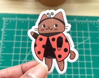 May include: A sticker of a brown cat dressed as a ladybug. The cat is wearing a red and black ladybug costume with black spots. The cat is smiling and has a friendly expression.