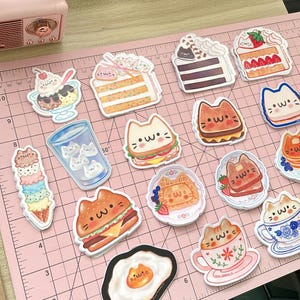 May include: A set of 14 colorful stickers featuring cute cartoon cats enjoying various foods and drinks. The stickers include a cat eating a sandwich, a cat drinking coffee, a cat eating a waffle, a cat eating a burger, a cat eating an egg, a cat eating a cake, a cat eating ice cream, a cat drinking a glass of water, and a cat eating a slice of cake. The stickers are perfect for adding a touch of whimsy to your laptop, water bottle, or planner.
