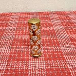 May include: A cylindrical lipstick tube with a metallic gold finish, featuring a repeating white floral pattern across its body. The top cap is also gold and displays the embossed text "JOY" on its flat surface.