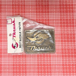 May include: A rectangular brass-toned metal belt buckle featuring a raised graphic of a bird head and the text Thrush. The buckle is enclosed in a clear plastic bag with an attached white paper tag displaying the Thrush logo and text #97179.