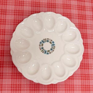 May include: A round white ceramic deviled egg serving platter featuring twelve oval-shaped indentations arranged in a circle. The center of the plate is decorated with a small circular pattern of blue and brown abstract geometric shapes.