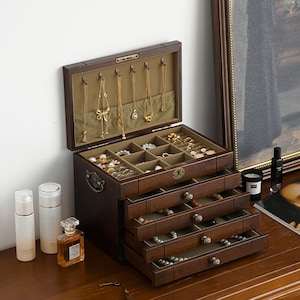 May include: A dark brown wooden jewelry box with multiple drawers and compartments. The top section is open, displaying necklaces hanging from hooks. The drawers are filled with various jewelry pieces. The box is on a wooden surface.