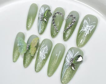 May include: A set of ten almond-shaped artificial nails. The nails are painted in a gradient of green with glitter at the tips. Some nails feature white butterfly designs, rhinestones, and 3D butterfly embellishments.