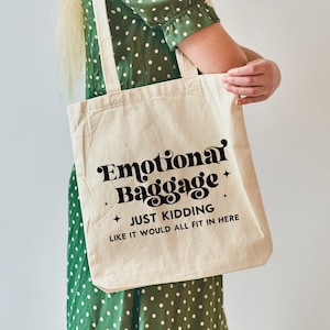 May include: A beige canvas tote bag featuring black text that reads Emotional Baggage, JUST KIDDING, LIKE IT WOULD ALL FIT IN HERE, accented with small four-pointed stars. The bag has two long shoulder straps and a rectangular, flat-bottomed design.