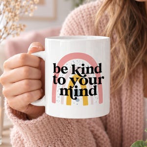 May include: A white ceramic mug featuring a graphic of a multi-colored rainbow in shades of pink, yellow, and blue with speckled details. The text "be kind to your mind" is printed in bold black lettering across the center of the rainbow design.
