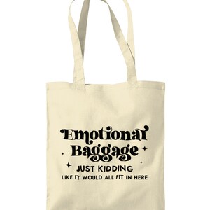 May include: A cream-colored canvas tote bag with two shoulder straps. The front features black text that reads Emotional Baggage, JUST KIDDING, LIKE IT WOULD ALL FIT IN HERE, accented by four small four-pointed star icons surrounding the text.
