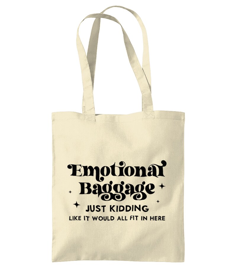 May include: A cream-colored canvas tote bag with two shoulder straps. The front features black text that reads Emotional Baggage, JUST KIDDING, LIKE IT WOULD ALL FIT IN HERE, accented by four small four-pointed star icons surrounding the text.