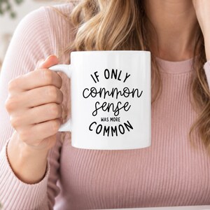 May include: A white ceramic mug featuring the black printed text IF ONLY common sense WAS MORE COMMON. The mug has a standard cylindrical shape with a curved handle on the side, displaying a clear contrast between the dark lettering and the white surface.